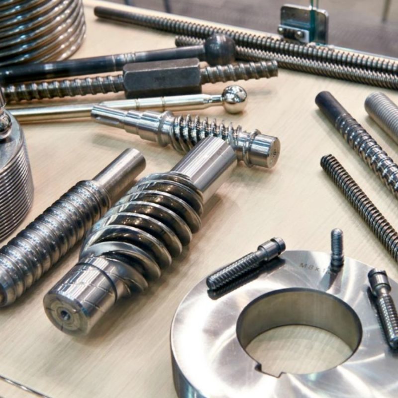 Various metallic industrial components, including screws, gears, and rings, displayed on a light-colored surface.