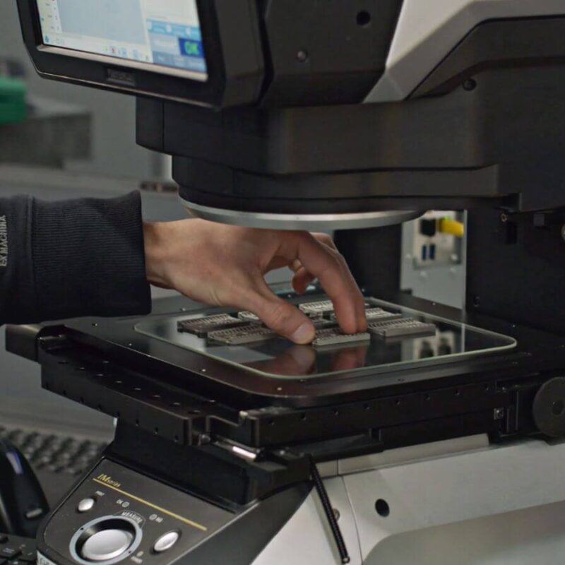 A person operates a machine by placing semiconductor wafers on a tray beneath a screen, in a lab or manufacturing setting.
