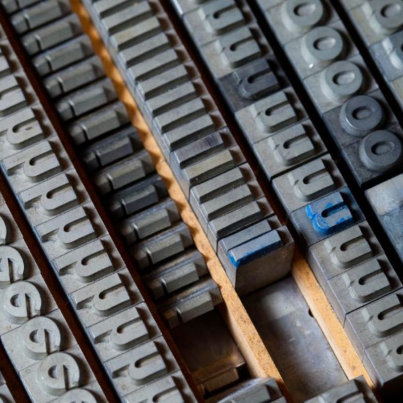 A close-up of metal letterpress type blocks arranged in rows, including letters, numbers, and symbols, typically used in traditional printmaking.