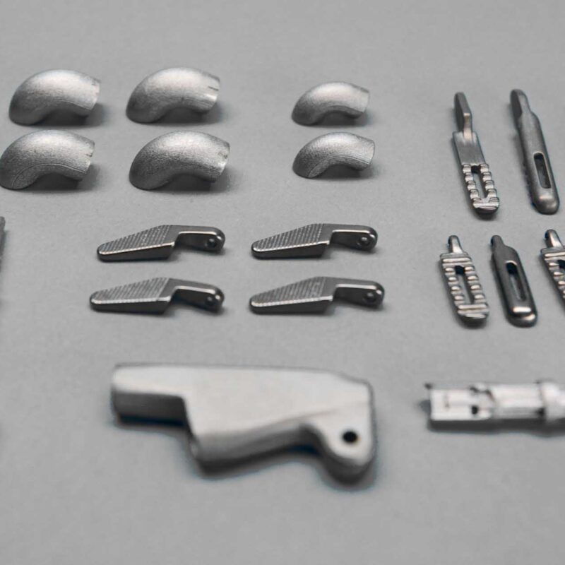 An assortment of metal components is neatly arranged on a gray surface, including curved pieces, connectors, and various small mechanical parts.