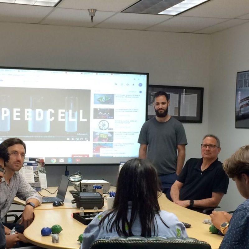 A group of people sit around a conference table during a meeting. A person stands near a screen displaying "SPEEDCELL." The room has posters and a whiteboard on the walls.