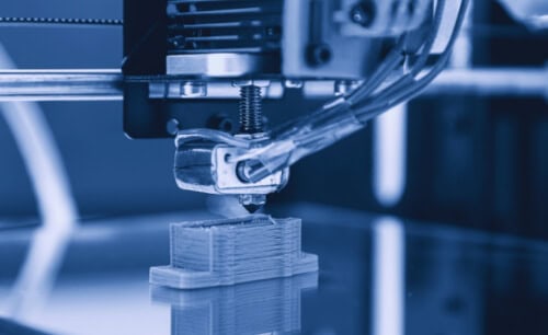 A close-up of a 3D printer in action, depositing a filament layer by layer to form a small rectangular object.