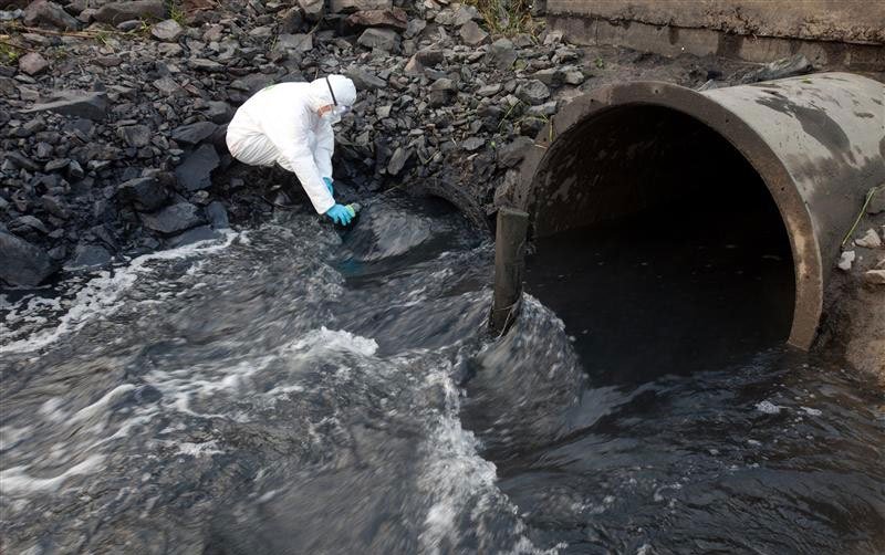 A person in protective clothing collects water samples from a stream flowing out of a large metal drainage pipe.