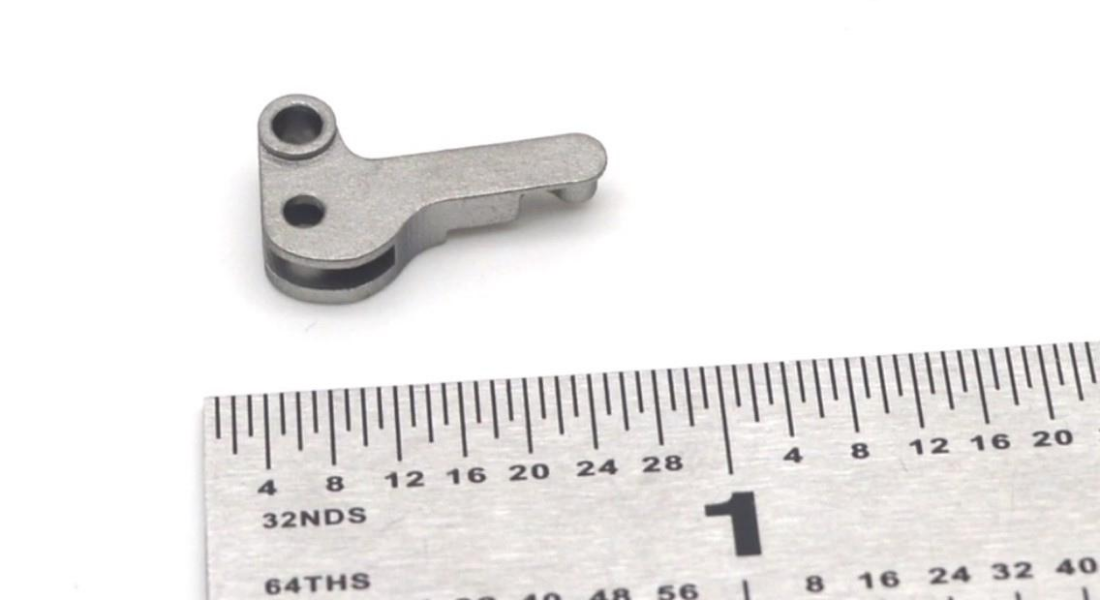 3DEO Wins MPIF Design Excellence Award Small metal component featuring intricate geometries, next to a ruler displaying measurements in inches, highlighting the precise size of the component.