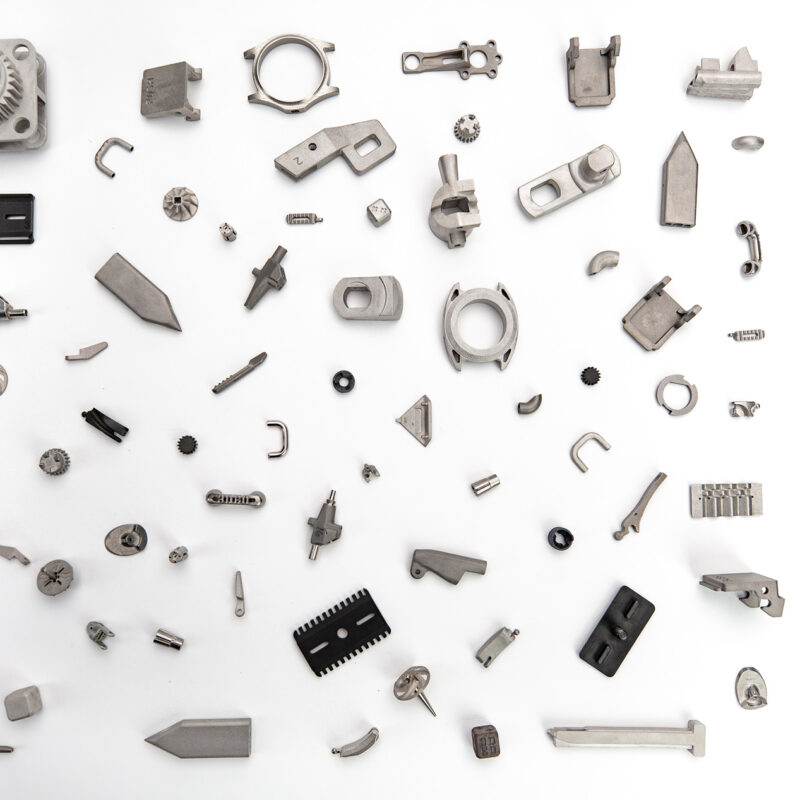 A variety of metal machine parts and components displayed on a white surface, arranged randomly. Items include gears, brackets, and other mechanical elements of different shapes and sizes.
