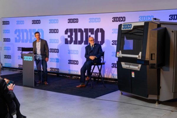 Two men, one standing at a podium and the other seated, at a presentation with a large 3d printer and an audience in a room with a logo-covered backdrop.