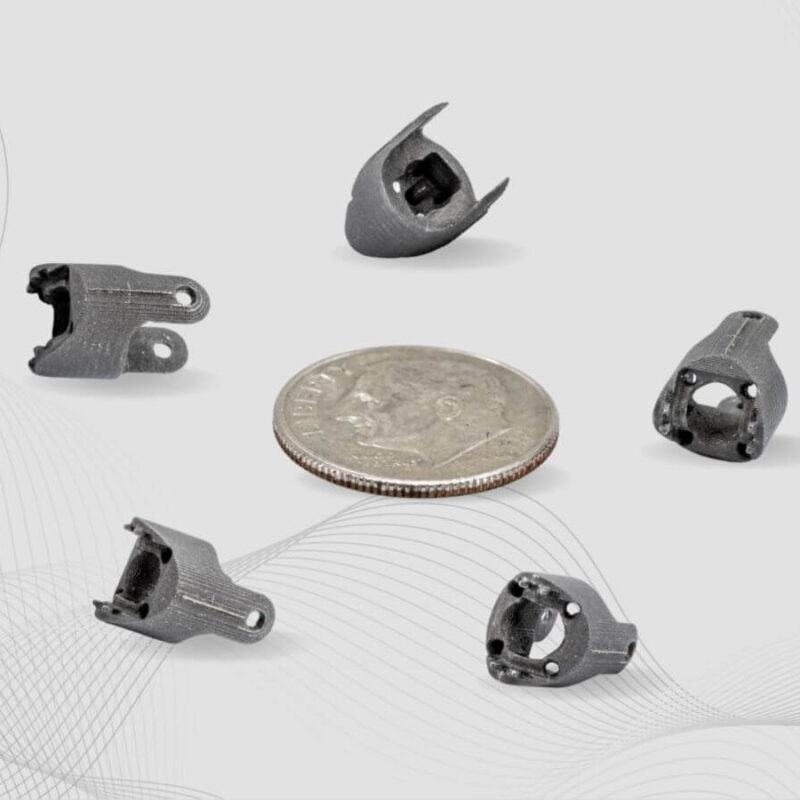 Five small metal 3D-printed parts, including a stainless steel clevis, are displayed around a quarter for size comparison. The background features a light, abstract wave pattern.