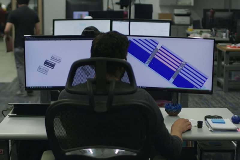 3DEO Adds Design for Additive Manufacturing (DfAM) as Core Service A person sits at a desk using a dual-monitor setup with various diagrams and data displayed on the screens in a modern office environment.