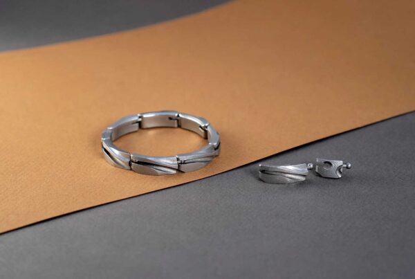 A silver bracelet and a pair of silver earrings, crafted through metal additive manufacturing, are placed on a brown and gray surface. The bracelet features a segmented design, while the earrings have a matching pattern.