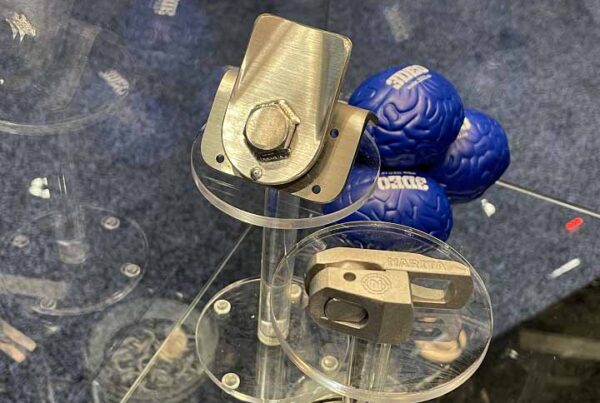 A display with two small metal mechanical components on clear stands, and multiple blue stress balls shaped like brain models in the background, highlights 3DEO talks pure copper and 3D printing for rail with 3DPrint.com.