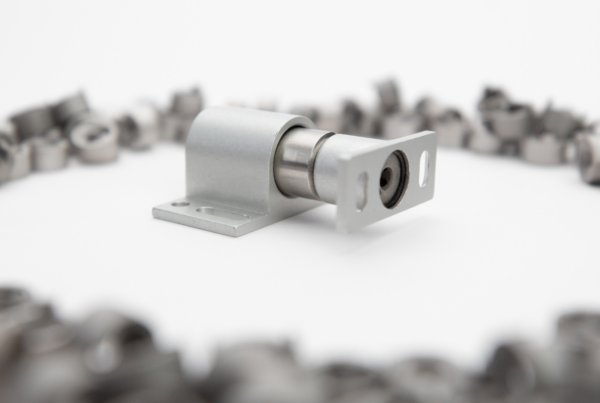 Metal sliding door roller hardware on a white surface, surrounded by blurred metal coils in the background, showcasing the ingenuity of US based manufacturing.